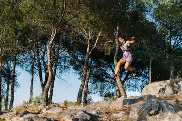 From below side view of active sportive young woman jumping high over rocky trail while running in green forest in mountains - ADSF17597