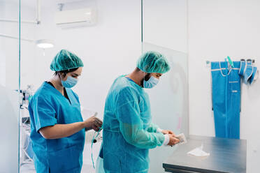 Seitenansicht von Tierärzten, die Schutzkleidung und Handschuhe anziehen, während sie im hellen Operationssaal eines Tierkrankenhauses stehen und sich auf eine Operation vorbereiten - ADSF17600