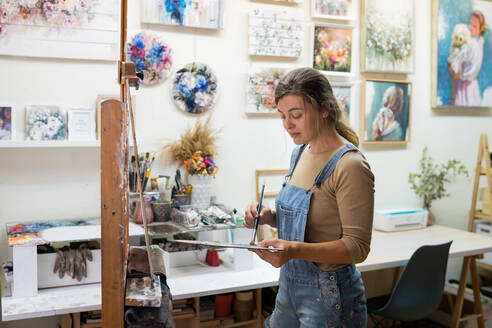 Seitenansicht einer Künstlerin, die an einer Staffelei steht und ein buntes Bild mit Farben malt, während sie in einer Kreativwerkstatt arbeitet - ADSF17603