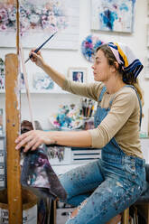 Seitenansicht einer Künstlerin, die an einer Staffelei sitzt und ein buntes Bild mit Farben malt, während sie in einer Kreativwerkstatt arbeitet - ADSF17605