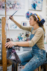 Side view of female artist sitting near easel and drawing colorful picture with paints while working in creative workshop - ADSF17605