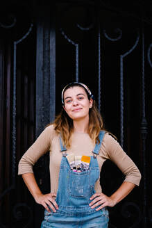 Fröhliche junge Künstlerin mit Pinseln und Farbtuben in der Tasche ihres Jeans-Overalls, die in die Kamera schaut, während sie in einer Werkstatt vor einem dunklen Hintergrund steht - ADSF17607