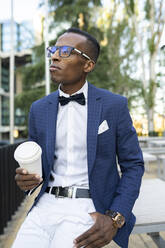 Zufriedener afroamerikanischer männlicher Unternehmer im eleganten Anzug, der mit einem Kaffee zum Mitnehmen auf der Straße sitzt und auf seinem Handy surft - ADSF17612