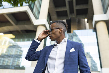Niedriger Winkel eines männlichen afroamerikanischen Unternehmers mit elegantem Anzug und Sonnenbrille, der in der Stadt steht und wegschaut - ADSF17613