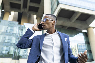 Von unten schwarzer männlicher Unternehmer in formellem Anzug und mit Sonnenbrille, der in der Nähe eines gläsernen Bürogebäudes steht und über ein Geschäftsprojekt am Mobiltelefon diskutiert, während er wegschaut - ADSF17615