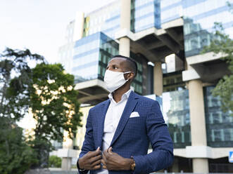 Seriöser schwarzer männlicher Unternehmer in formellem Anzug und medizinischer Maske steht in der Innenstadt und wartet auf ein Treffen und schaut weg - ADSF17617