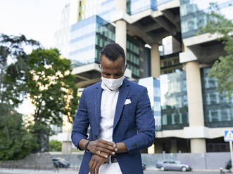 Niedriger Blickwinkel auf einen seriösen schwarzen männlichen Unternehmer in einem formellen Anzug und einer medizinischen Maske, der in der Innenstadt steht und die Zeit auf seiner Armbanduhr überprüft, während er auf ein Treffen wartet - ADSF17618