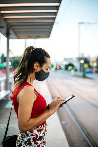 Side view of young female in black protective mask browsing smartphone while standing on railway platform and waiting for train stock photo