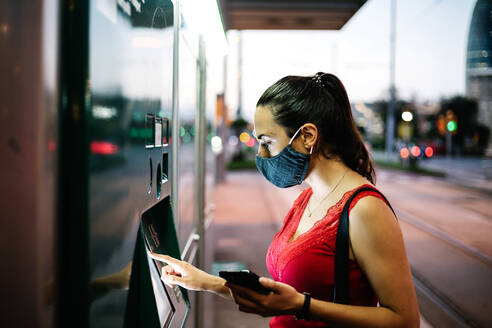 Seitenansicht eines jungen weiblichen Fahrgastes mit Schutzmaske und Mobiltelefon, der an einem Automaten eine Fahrkarte für öffentliche Verkehrsmittel kauft - ADSF17635