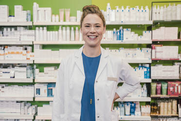 Happy pharmacist with hand on hip standing in chemist shop - MFF06798