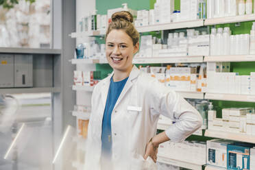Happy pharmacist with hand on hip standing in chemist shop - MFF06799