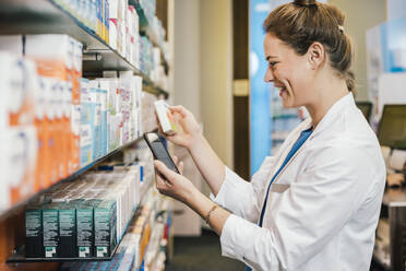 Happy pharmacist with smart phone checking medicine while working at pharmacy - MFF06804