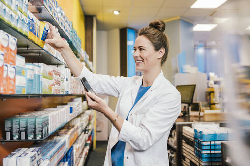 Lächelnde Apothekerin mit Smartphone bei der Überprüfung von Medikamenten in einer Apotheke - MFF06805