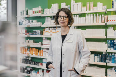 Confident pharmacist with hands in pockets standing in chemist shop - MFF06806