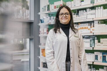 Confident pharmacist wearing lab coat while standing in chemist shop - MFF06822