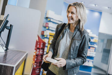 Smiling female customer with medicine standing at checkout counter in chemist shop - MFF06824