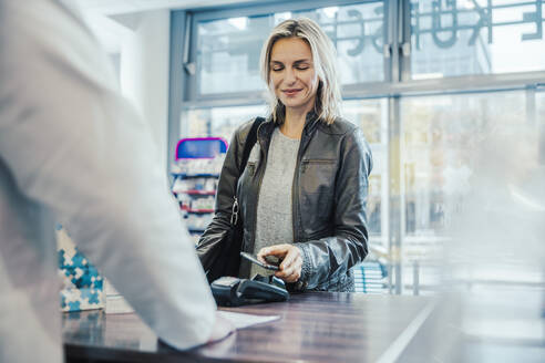 Lächelnder Kunde beim Bezahlen mit dem Handy an der Kasse eines Drogeriemarktes - MFF06829