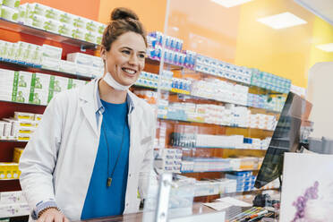 Confident pharmacist with surgical mask standing at checkout counter in chemist shop - MFF06862