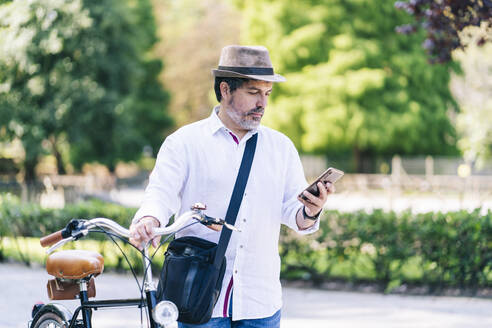 Älterer Mann, der ein Mobiltelefon benutzt, während er mit dem Fahrrad in einem öffentlichen Park steht - DGOF01644