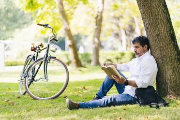 Mature man reading book while leaning on tree trunk at public park - DGOF01650