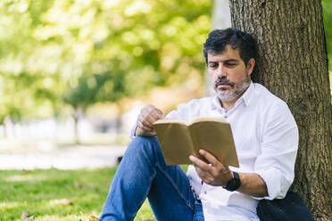 Mature man reading book while leaning on tree trunk in public park - DGOF01651