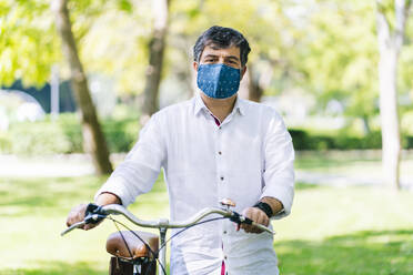 Mature man with protective face mask with bicycle at public park during COVID-19 - DGOF01653