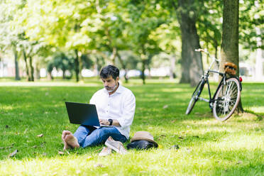 Älterer Mann, der einen Laptop benutzt, während er im öffentlichen Park im Gras sitzt - DGOF01655