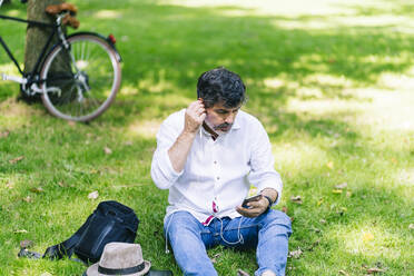 Mature man listening to music through earphones while sitting on grass at public park - DGOF01657