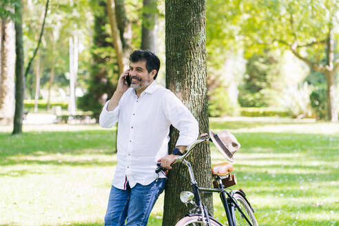 Happy mature man talking on smart phone while leaning on tree at public park - DGOF01663