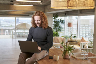 Smiling redhead man using laptop sitting on table at home - FMKF06663