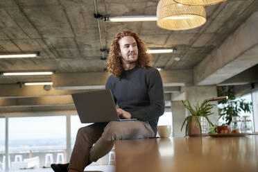 Thoughtful redhead man looking away while using laptop sitting at home - FMKF06664