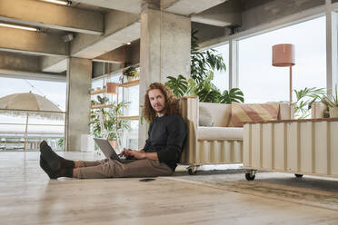 Redhead man using laptop sitting on floor in living room at home - FMKF06668