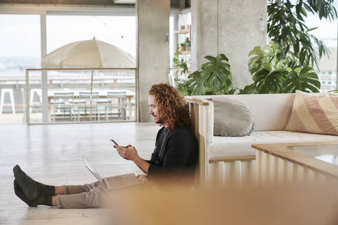 Mittlerer erwachsener Mann, der ein Smartphone benutzt und mit einem Laptop auf dem Boden im Wohnzimmer sitzt - FMKF06671