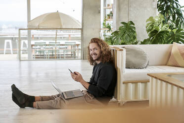 Smiling man using smart phone sitting with laptop on floor in living room - FMKF06672