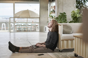 Redhead man looking up while using laptop while sitting in living room at home - FMKF06675