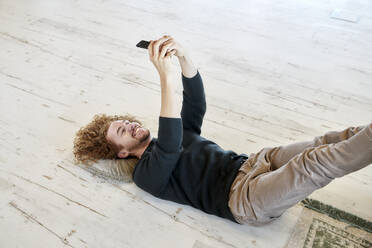 Smiling man taking selfie through smart phone lying down on floor at home - FMKF06677