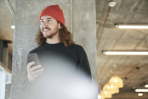 Man wearing red hat using smart phone standing against column at home stock photo