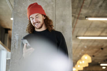 Mann mit rotem Hut, der mit seinem Smartphone Textnachrichten verschickt, steht an einer Säule zu Hause - FMKF06682