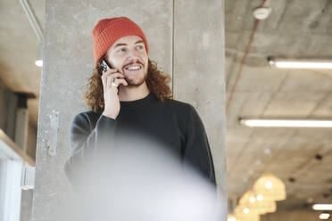 Mann mit rotem Hut, der sein Smartphone aufnimmt, steht an einer Betonsäule zu Hause - FMKF06683