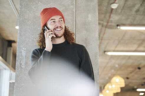 Man wearing red hat taking on smart phone standing against concrete column at home - FMKF06683