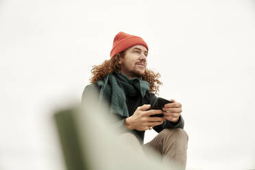 Nachdenklicher Mann, der sein Smartphone benutzt und wegschaut, während er auf der Terrasse eines Gebäudes gegen den klaren Himmel sitzt - FMKF06691