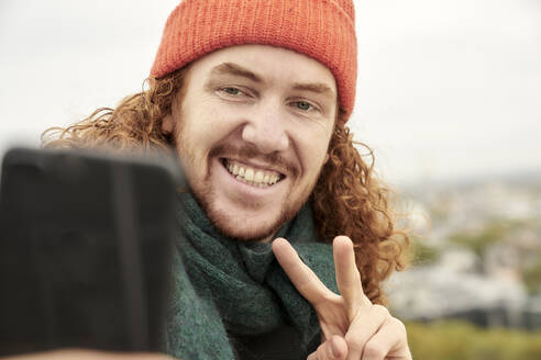 Smiling man doing peace sign while taking selfie on smart phone - FMKF06693