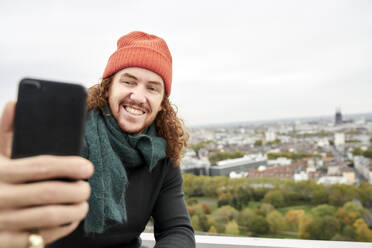 Man smiling while taking selfie through smart phone on building terrace against sky - FMKF06694