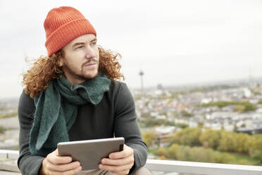 Mann schaut weg, während er ein digitales Tablet benutzt, das auf einer Gebäudeterrasse gegen den Himmel sitzt - FMKF06696