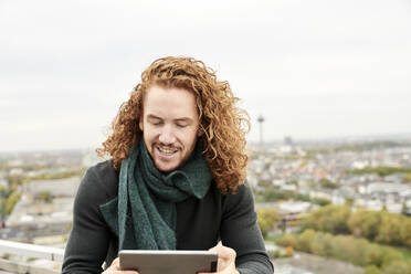 Rothaariger Mann, der ein digitales Tablet benutzt, während er auf der Terrasse eines Gebäudes gegen den Himmel sitzt - FMKF06697
