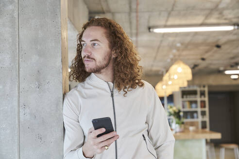 Hipster man looking away while holding smart phone leaning on concrete column at home - FMKF06701