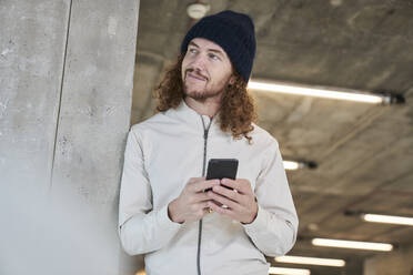 Lächelnder Hipster-Mann mit Strickmütze, der sein Smartphone benutzt, während er sich zu Hause an eine Säule lehnt - FMKF06706