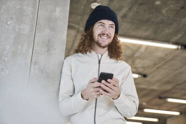 Hipster-Mann schaut weg und benutzt sein Smartphone, während er sich zu Hause an eine architektonische Säule lehnt - FMKF06707