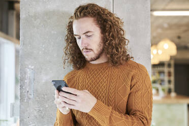 Hipster Mann Textnachrichten auf Smartphone stehend gegen graue Säule im Wohnzimmer zu Hause - FMKF06714