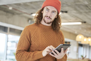 Hipster-Mann, der ein digitales Tablet benutzt und wegschaut, während er zu Hause im Wohnzimmer steht - FMKF06718